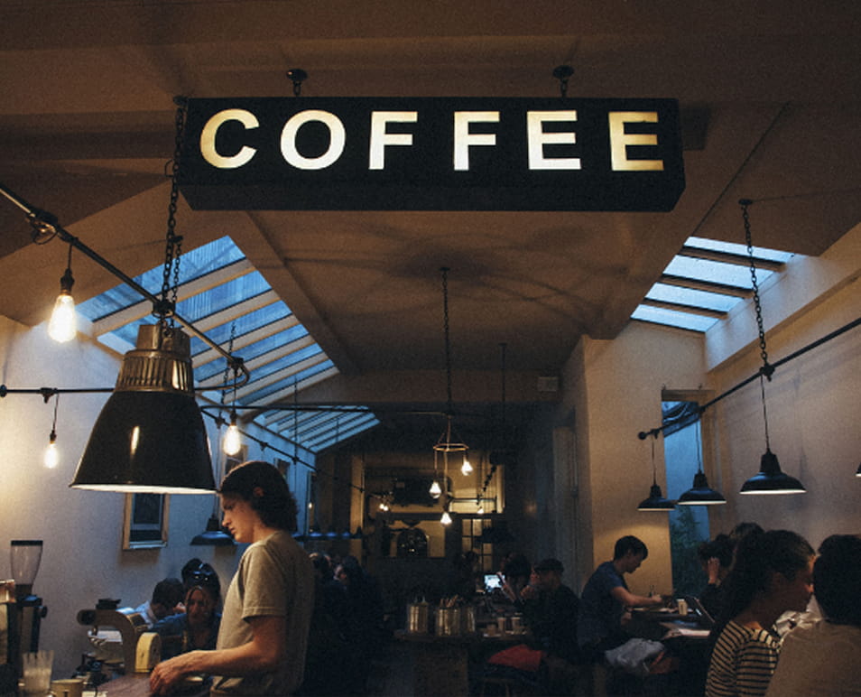 Modern coffee shop interior with an illuminated COFFEE sign, industrial lighting, barista at the counter, and seated guests