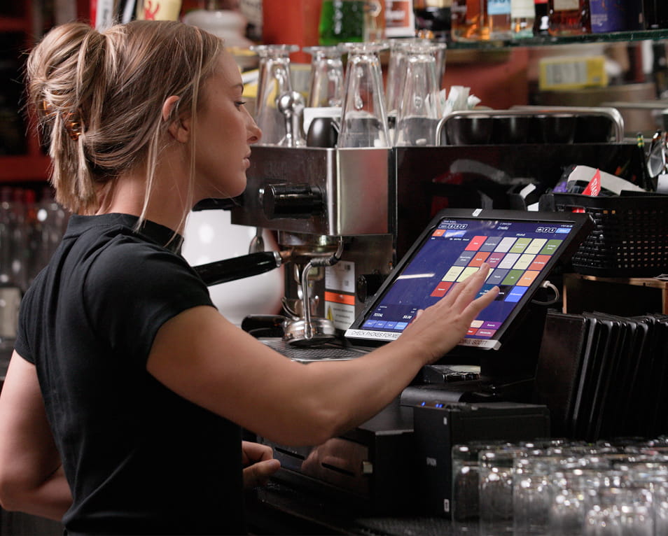 Bartender using a touchscreen tablet POS and receipt printer behind a bar with glassware and bottles