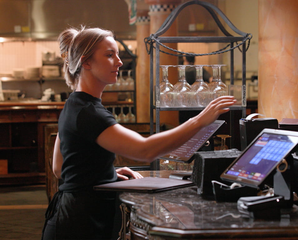 Server using a tablet POS on a restaurant counter with a receipt printer and open kitchen in the background